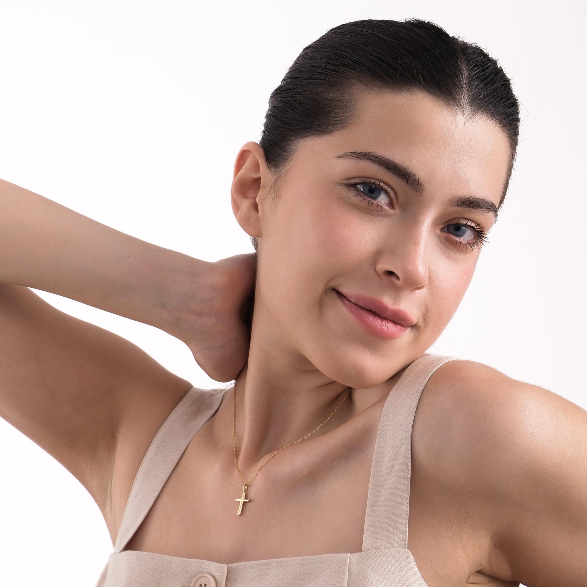 Smiling woman wearing a 14K solid gold cross necklace, styled with a beige buttoned top, hand resting behind her neck.