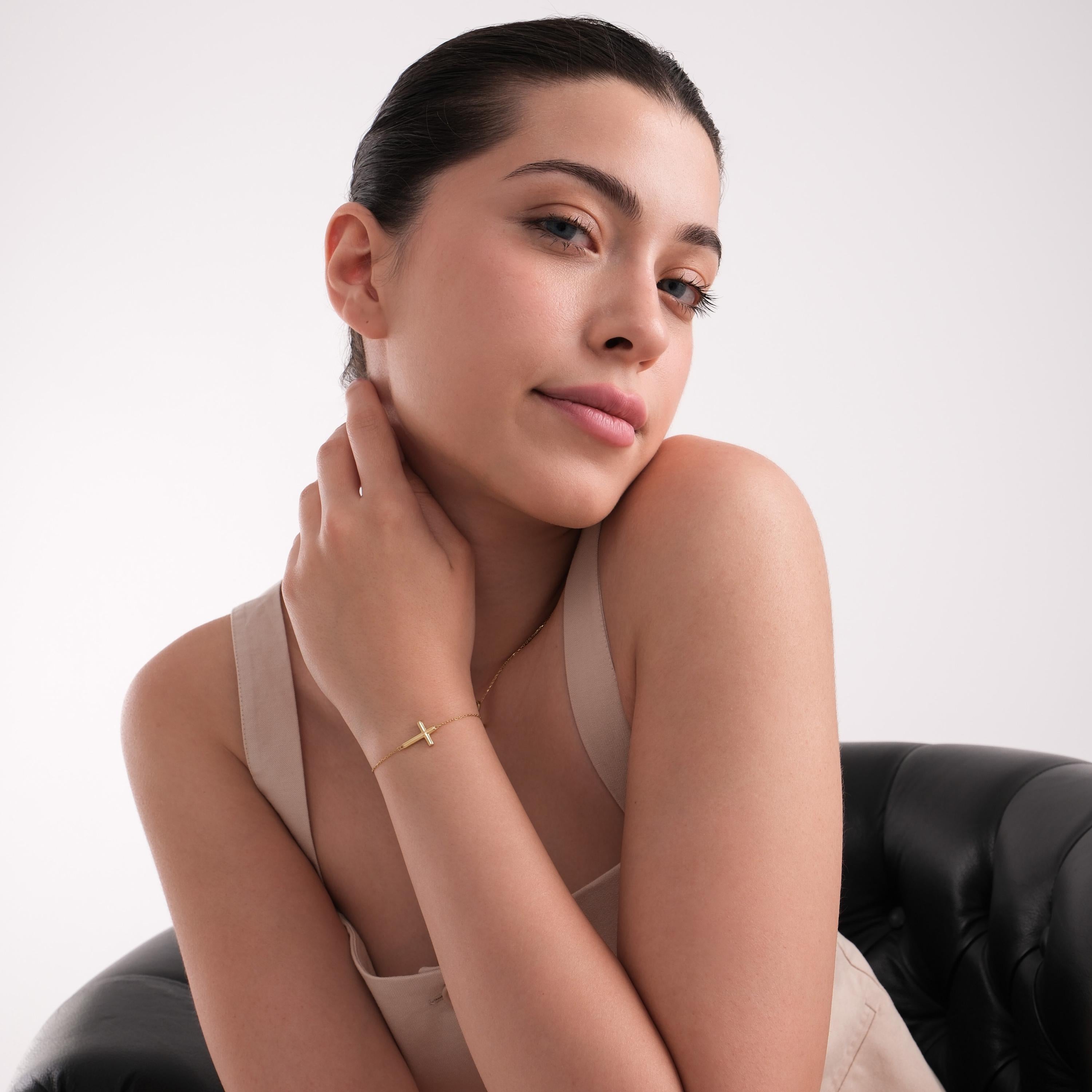 Stylish woman posing in neutral tones, wearing a 14K solid gold dainty cross bracelet with a chunky horizontal cross centerpiece.