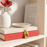 18K Gold Vermeil Custom Picture Necklace displayed on a white ceramic dish in front of stacked red and pink books and a white vase with flowers, engraved family portrait pendant