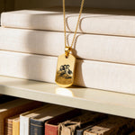 18K Gold Vermeil Custom Picture Necklace featuring a rectangular dog tag pendant engraved with a family portrait, displayed on a gold chain over stacked neutral books on a shelf