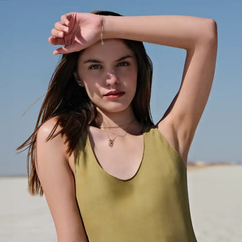 A young woman stands outdoors with her left arm raised to shield her eyes from the sun. She has long brown hair and wears a sleeveless green top. She accessorizes with two gold necklaces, one featuring a small pendant, and a delicate gold rosary bracelet on her left wrist. The background is a bright, open area under a clear blue sky.