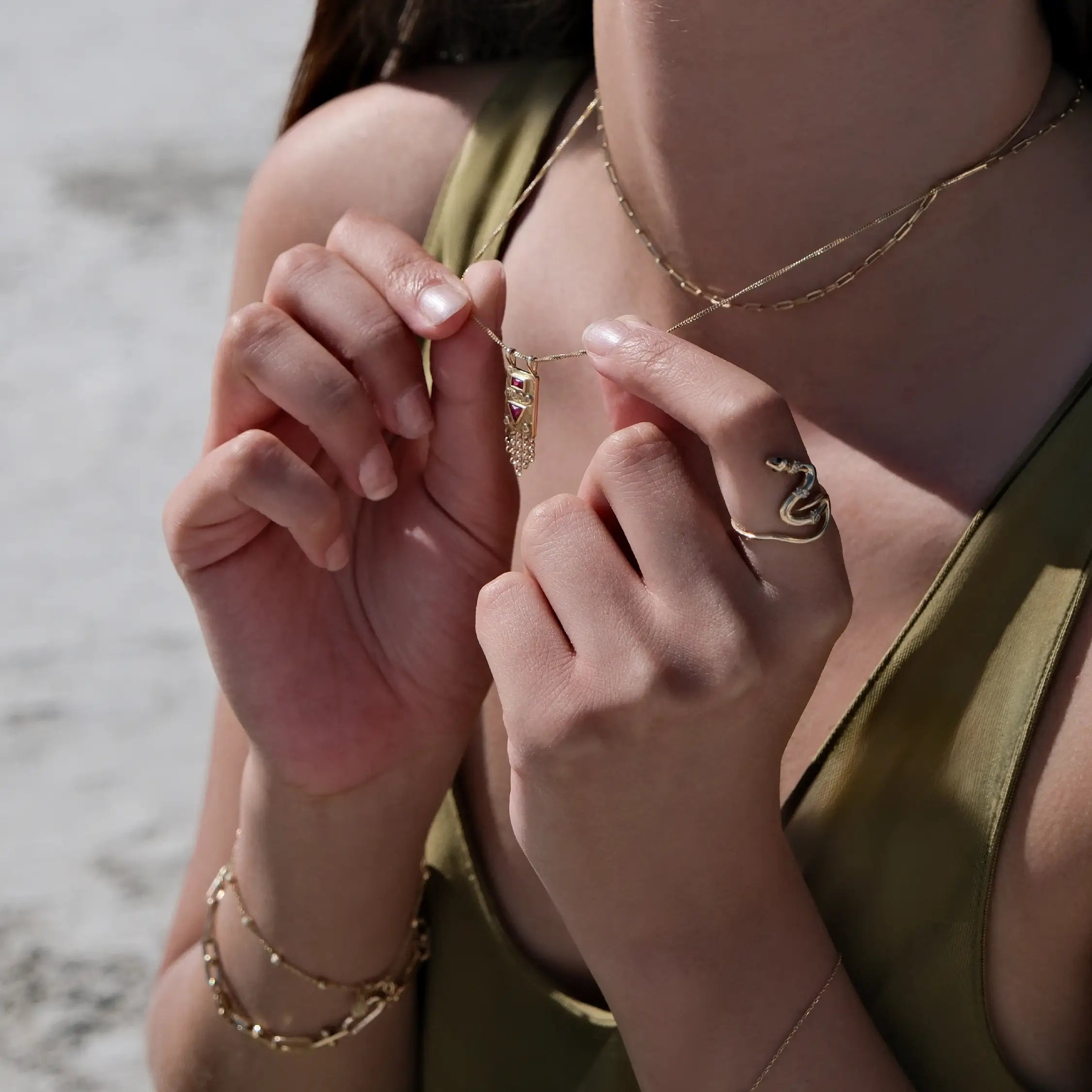 A woman wearing a green top holds a delicate necklace with a small pendant between her fingers. She has a gold rosary bracelet on her right wrist and wears a snake-shaped ring on her right index finger. Her background is an outdoor, sunlit area with a light-colored surface.