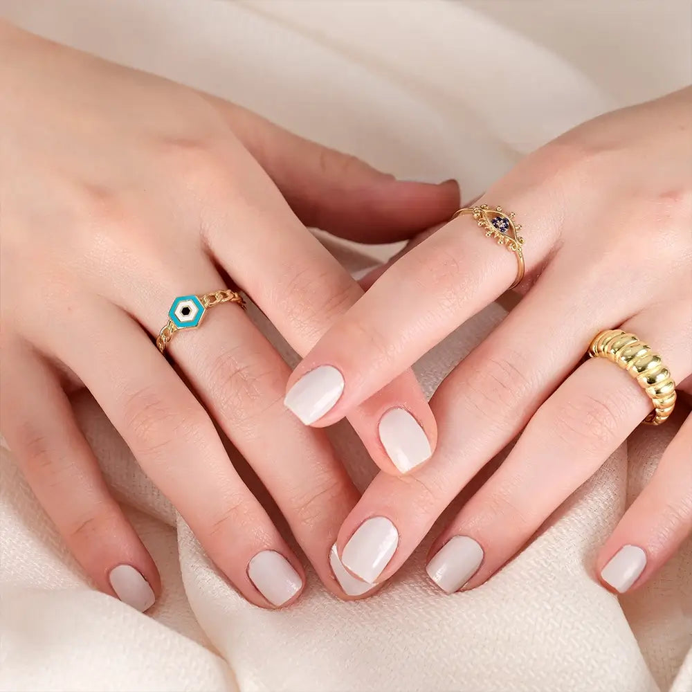 A close-up of a woman's hands adorned with various rings. Her left hand wears a gold ring with a hexagonal turquoise and white design on her index finger, while her right hand features a delicate gold ring with a central blue stone and small surrounding diamonds on her middle finger, and a thick gold ribbed band on her pinky finger. Her nails are neatly manicured with a light beige polish. The background is a soft, light-colored fabric.