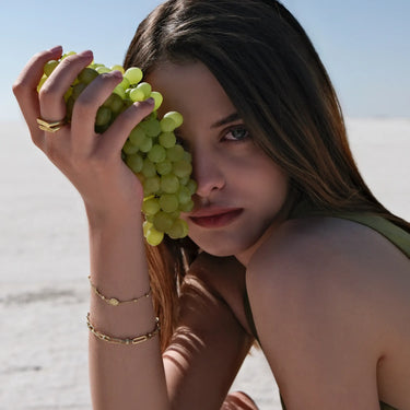 A young woman with long brown hair holds a bunch of green grapes against the left side of her face. She wears two gold bracelets on her right wrist, including a delicate gold rosary bracelet. Her expression is contemplative, and she is set against a minimalist, bright outdoor background.